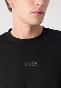 T-shirt noir à col rond, avec un logo "BOSS" noir mat sur la poitrine. Le matériau semble lisse et ajusté.