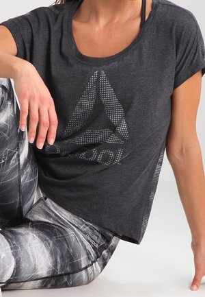 Kobieta w ciemnoszarej koszulce Reebok i czarno-białych legginsach we wzory, siedząca z podniesionym jednym kolanem i ręką opartą o nie.
