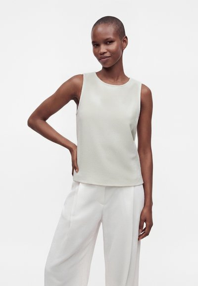 Personne aux cheveux courts pose avec assurance, portant un haut clair sans manches et un pantalon blanc ample sur un fond blanc uni.