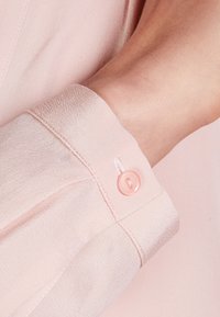 Tissu de blouse rose avec une texture lisse, présentant un détail de bouton sur le poignet. Le bouton est d'une couleur rose clair assortie.