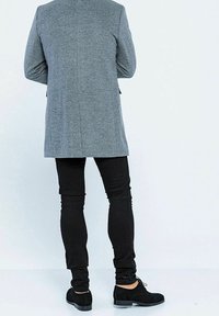 Cappotto grigio in misto lana con taglio sartoriale, abbinato a jeans neri slim-fit e scarpe stringate in suede nero, visto da dietro.
