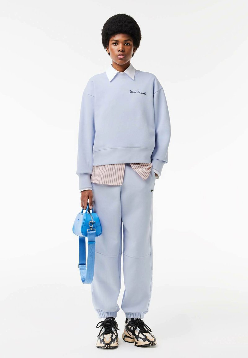 Sudadera azul clara con logo bordado, superpuesta a una camiseta de rayas rosas. Pantalones de chándal azules a juego y un pequeño bolso azul. Zapatillas en blanco y negro.