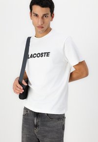Balta medvilnė marškinėliai su juodu "LACOSTE" logotipu. Apvalus iškirpimas, trumpomis rankovėmis ir subtilus siuvinėtas logotipas apačioje.