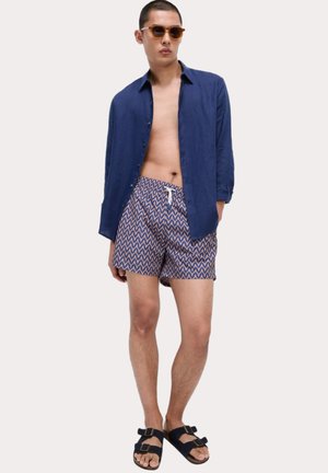 Marineblaues Leinenhemd, gemusterte Badeshorts mit blauem und orangefarbenem Zickzackdesign, braune Sonnenbrille und schwarze Sandalen mit zwei Schnallen.
