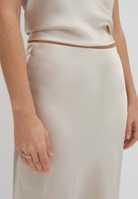 Jupe texturée couleur crème avec un subtil éclat, présentant un design ajusté et une taille lisse. Main reposant sur l'ourlet.