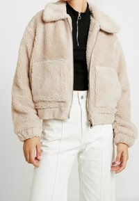 Beige kort faux fur-jacka med krage, två framfickor och dragkedjestängning. Bärs över en svart topp och vita byxor.