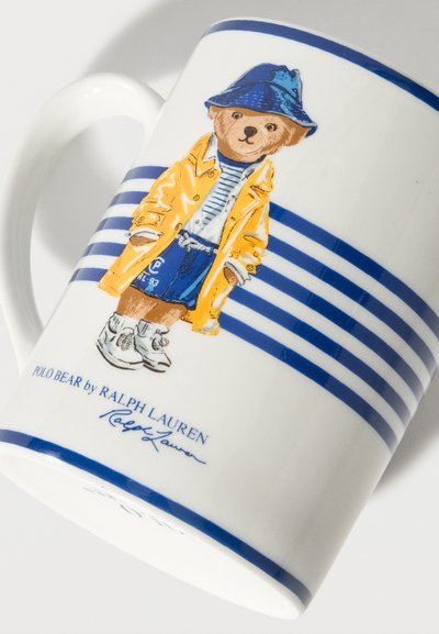 Fehér kerámia bögre kék csíkokkal és egy medvével, aki sárga esőkabátot, kék sapkát, csíkos inget, kék rövidnadrágot és fehér tornacipőt visel.