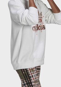 Vit hoodie med en blommig röd och svart "adidas"-logotyp. Visar en avslappnad passform. I kombination med byxor i rutigt mönster i varma nyanser.