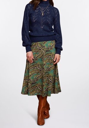 Frau trägt navyblauen Strickpullover, grün-braun gemusterten Midirock, braune Lederstiefel, goldene Halsketten und Ringe, steht vor weißem Hintergrund.