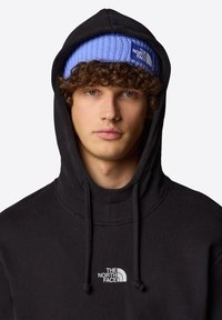 Felpa nera con cappuccio, cordini e logo bianco. Abbinata a un berretto di maglia azzurro chiaro con etichetta del marchio.