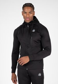 Zwarte sportieve zip-up hoodie met hoge kraag, twee voorzakken en een logo op de borst. Glad materiaal met een getailleerd ontwerp en lange mouwen.