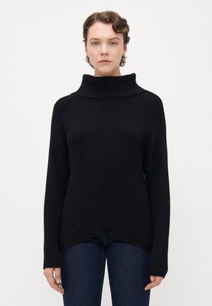 En person som har på seg en svart ribbestrikket høyhalset genser og mørkeblå jeans, står mot en ensfarget hvit bakgrunn.