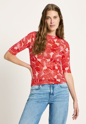 Woman with long wavy hair wears red floral patterned top with elbow-length sleeves and light blue jeans, posing with one hand on hip.