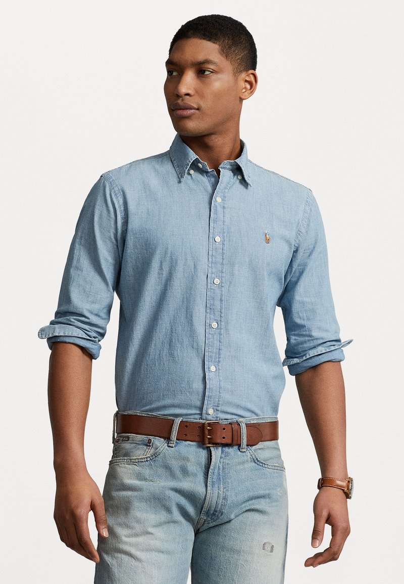 Light blue chambray shirt with button collar, rolled-up sleeves, and small logo on the left chest; paired with faded denim and brown leather belt.