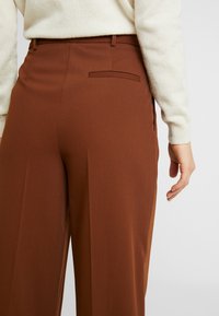 Bruna byxor med vida ben och subtil struktur, med en enda bakficka och en rak midjeresår. Bärs med en ljus tröja.