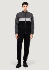 Chaqueta de cremallera en negro y gris con un diseño de bloques de color, combinada con pantalones negros. La chaqueta tiene un cuello alto y bolsillos en el frente.