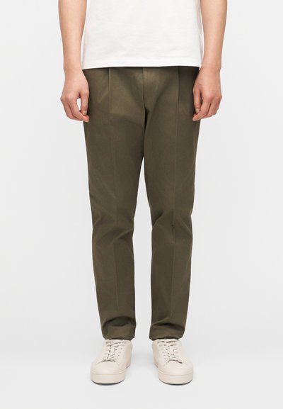 Personne portant un pantalon droit vert olive, des baskets blanches et une chemise blanche, se tenant devant un fond clair uni.