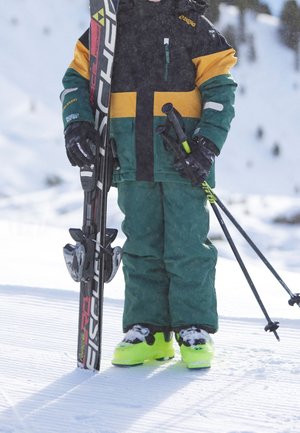 Enfant en tenue de ski verte et jaune tenant des skis et des bâtons sur une pente enneigée avec des chaussures de ski vert néon.
