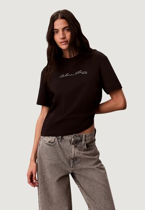 Kobieta o długich ciemnych włosach, ubrana w czarny T-shirt Calvin Klein i jasnoszare dżinsy, stojąca na neutralnym tle.