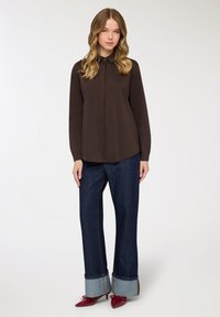 Camicia marrone a maniche lunghe con bottoni e colletto a punta, abbinata a pantaloni denim scuri a gamba larga con risvolti e scarpe rosse con tacco.