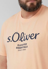 Perzikkleurig katoenen T-shirt met korte mouwen, ronde hals en marineblauwe tekstprint met de tekst "s.Oliver ELEVATED ESSENTIALS SINCE 1969".