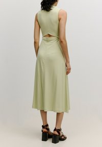 Hellgrünes, ärmelloses Kleid mit einer taillierten Oberteile und ausgeschnittenem Rücken. Der ausgestellte Rock reicht bis zur Mitte der Wade und wird mit dunkelbraunen Blockabsatzsandalen kombiniert.