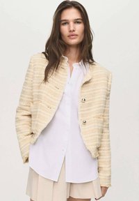 Cream and beige textured cropped jacket with two silver buttons, worn over a white button-up shirt and light beige skirt.