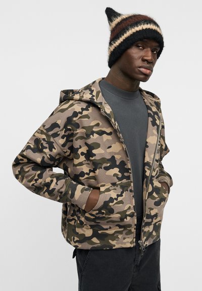 Φούτερ με φερμουάρ και καμουφλάζ σε αποχρώσεις καφέ, πράσινου και μαύρου. Διαθέτει μπροστινή τσέπη και κουκούλα. Συνδυάζεται με πλεκτό σκουφάκι σε μαύρο, καφέ και κρεμ.