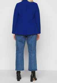 Chaqueta azul royal con un diseño entallado, abertura simple en la parte trasera, combinada con jeans de pierna recta de lavado medio y botines negros de tacón.