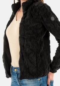 Chaqueta de piel sintética negra con cierre de cremallera en el frente, patrón texturizado, cuello alto y acento de logo en la manga. Combinada con una blusa clara y jeans azules.