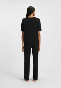 Vrouw met gevlochten haar die blootsvoets staat, een losse zwarte top met korte mouwen en zwarte wijde broek draagt, zich omkerend tegen een effen achtergrond.