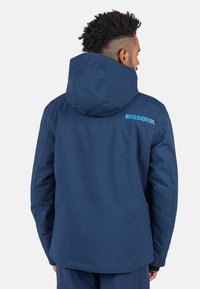 Marineblaue Outdoor-Jacke mit Kapuze, mit einer glatten Textur und dem "ROSSIGNOL"-Logo in Hellblau auf dem Rücken.