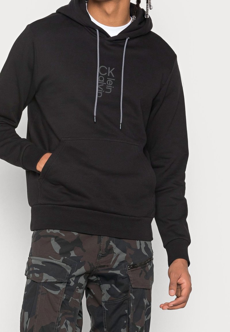 Felpa nera con cappuccio e tasca frontale, con lacci grigi e dettagli con logo. Abbinata a pantaloni cargo mimetici.