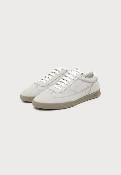 Paire de baskets basses blanches avec semelles en caoutchouc beige, lacets blancs et détails de couture subtils sur un fond clair uni.
