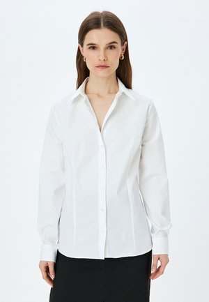 Camisa blanca de algodón con botones. Presenta un collar clásico, mangas largas con puños abotonados y un diseño ajustado.