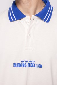 Krema polo majica z modrim prugastim ovratnikom, dva gumba na zapenjanju in vezenim napisom "KAOTIKO SOCIETY BURNING REBELLION" v modri barvi.