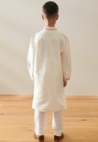 Weißes traditionelles Outfit mit einer langen Tunika mit bestickten Manschetten und passenden Hosen, getragen mit braunen Schuhen.
