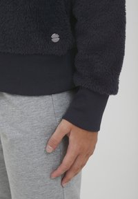 Meriino fleecetakki laivastonsinisellä värillä, jossa on ribatut hihansuut ja helma, ja hopeinen logo-tyyli. Yhdistetty harmaisiin teksturoituihin verkkareihin.