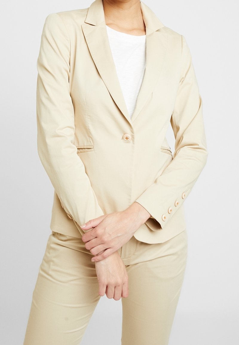 Blazer beige en tissu léger avec fermeture à un bouton, revers crantés et manches longues, doté de boutons décoratifs aux poignets.