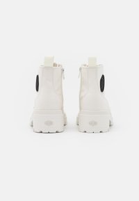 Bottines en toile blanche avec une semelle en caoutchouc texturée, une fermeture éclair latérale et un patch logo circulaire noir sur le côté extérieur.