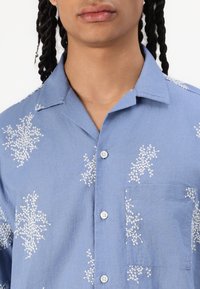 Chemise bleu clair en tissu avec broderies florales blanches. Présente un devant à boutons, une poche poitrine et un col.