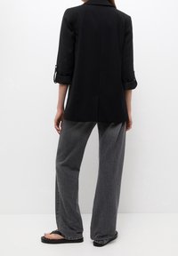 Blazer noir sur mesure avec manches trois-quarts, associé à un jean denim gris ample et des tongs noires. Design épuré et minimaliste.