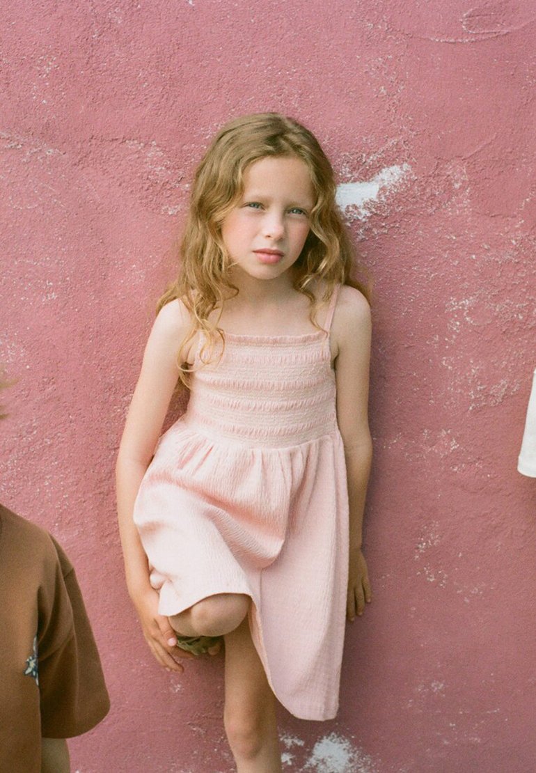Fille aux longs cheveux bouclés portant une robe rose clair se tient contre un mur rose texturé, genou plié.