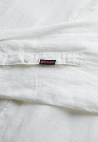 Camisa de lino blanca con cierre de botones y una etiqueta con la marca "SUPERDRY" en naranja sobre un fondo negro, destacando la textura de la tela.