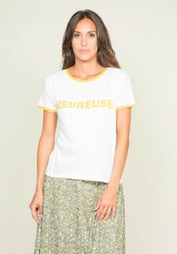 T-shirt en coton blanc avec des accents jaunes et un texte en gras "LEUREUSE" sur le devant. Associé à une jupe florale verte à motifs.
