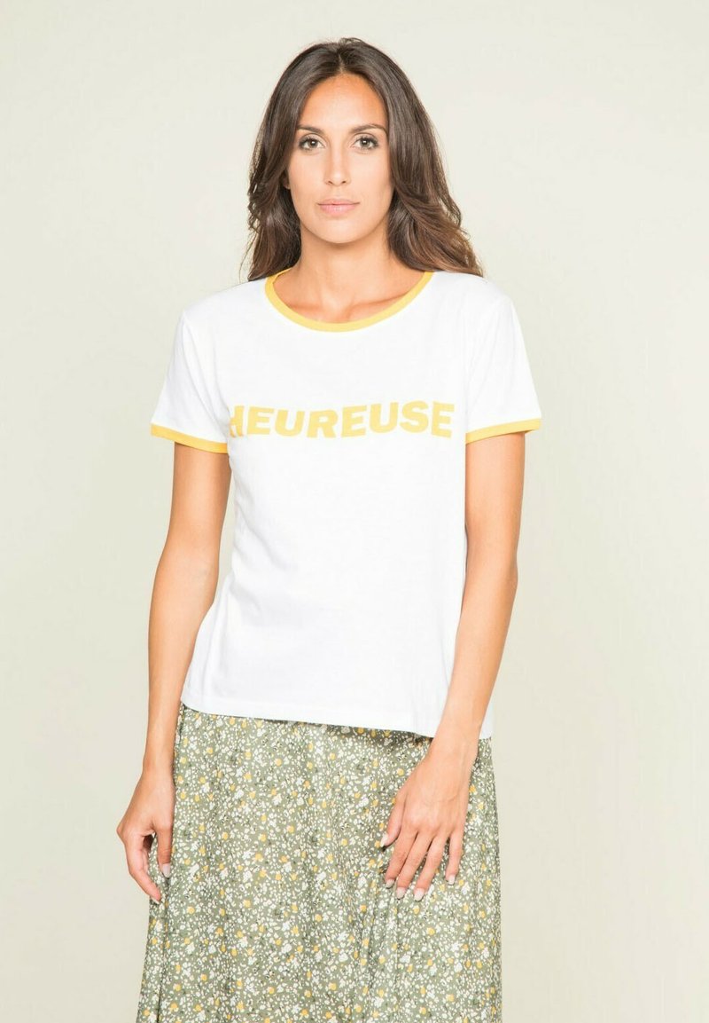 T-shirt en coton blanc avec des accents jaunes et un texte en gras "LEUREUSE" sur le devant. Associé à une jupe florale verte à motifs.