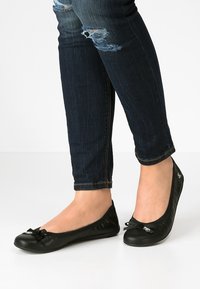 Svarta platta skor med rund tå, mjukt lädermaterial och en liten rosettdetalj fram. Bärs med mörka jeans.