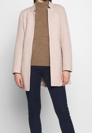 Person trägt einen hellbeigen offenen Mantel, braunen gestrickten Rollkragenpullover und dunkelblaue Slim-Fit-Hosen vor einem schlichten hellen Hintergrund.