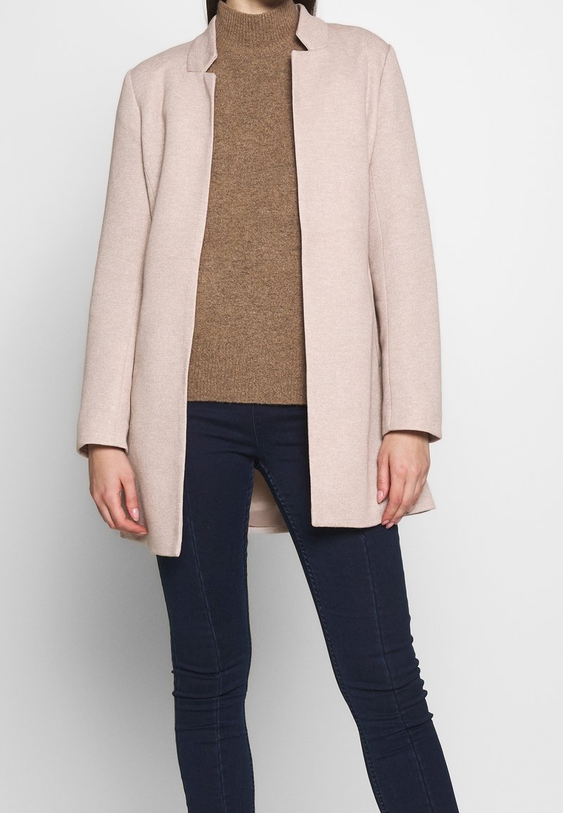 Personne portant un manteau ouvert beige clair, un pull à col roulé en laine marron et un pantalon slim bleu foncé sur un fond clair uni.