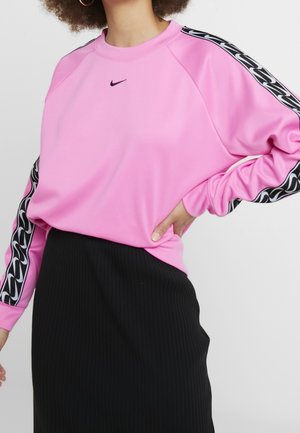 Kobieta w różowym swetrze Nike z czarno-białymi wzorzystymi paskami na rękawach i czarnej, prążkowanej spódnicy, ręka na biodrze.